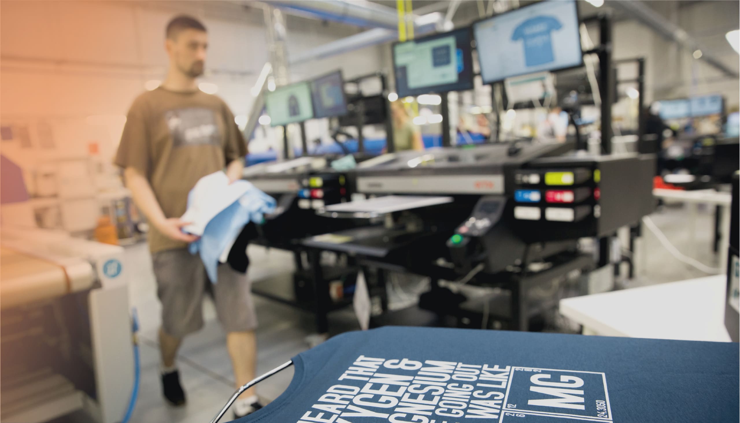 Foto. Im unteren Bildbereich liegt im Vordergrund ein blaues T-Shirt glatt aufgespannt. Im Hintergrund ist unscharf eine große Textildruckmaschine und ein Mitarbeiter zu sehen.